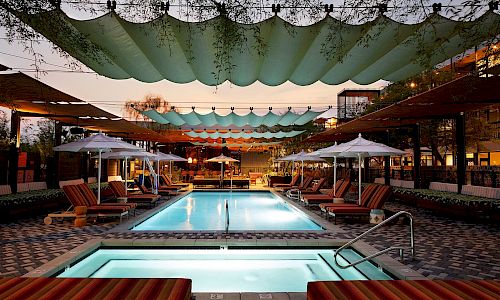 A stylish pool area with two turquoise-lit pools, lounge chairs, umbrellas, and decorative sails overhead at dusk, inviting relaxation.
