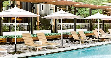 A resort pool area with sun loungers, white umbrellas, shaded seating, and wooden decking beside a clear blue pool, hotel-like and relaxing.