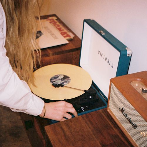 A person is placing a vinyl record on a turntable next to a Marshall speaker, while other records are on the table.