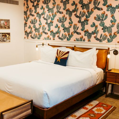 A cozy bedroom with a cactus-themed wallpaper, white bedding, a wooden bed frame, a colorful rug, wall art, and two nightstands with lamps.