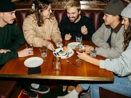 A group of five people is sitting around a table at a restaurant, sharing food and drinks, smiling and enjoying each other's company.