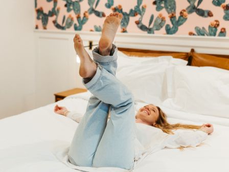 A person lying on a bed with their legs playfully raised, set against a colorful wall with a cactus pattern.