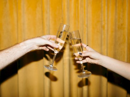 Two hands, one with nail polish, clink champagne glasses with a golden backdrop, suggesting a celebratory moment.