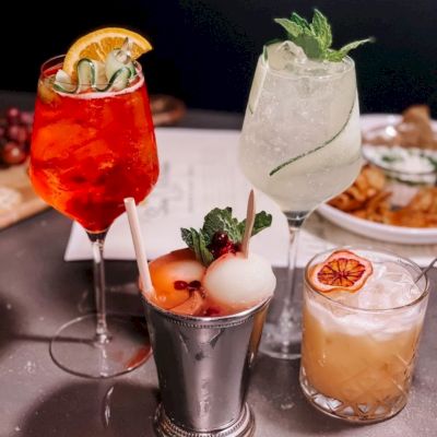 The image shows a table with four stylish cocktails in different glasses, garnished with fruit and herbs. A plate of fries is in the background.