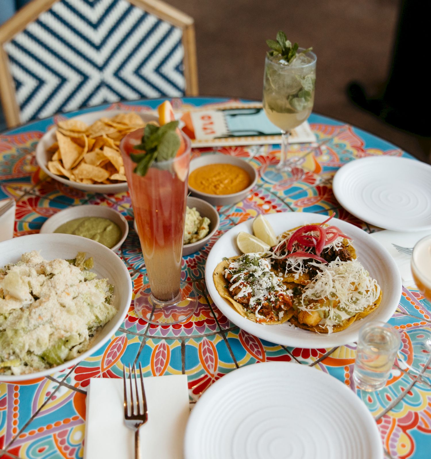 A colorful table with a variety of dishes including salads, chips, and drinks, set with empty plates and napkins.