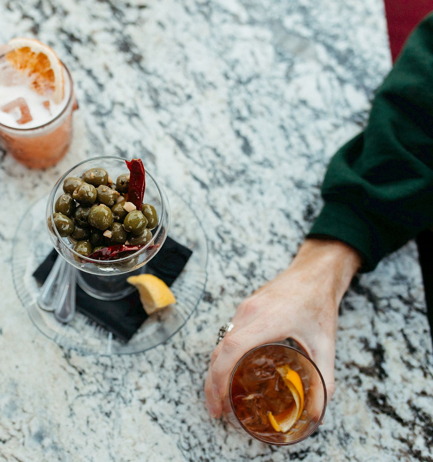 The image shows two people sitting at a table with drinks and a small dish of olives. One is holding a cocktail with ice, the other a drink with a citrus slice.