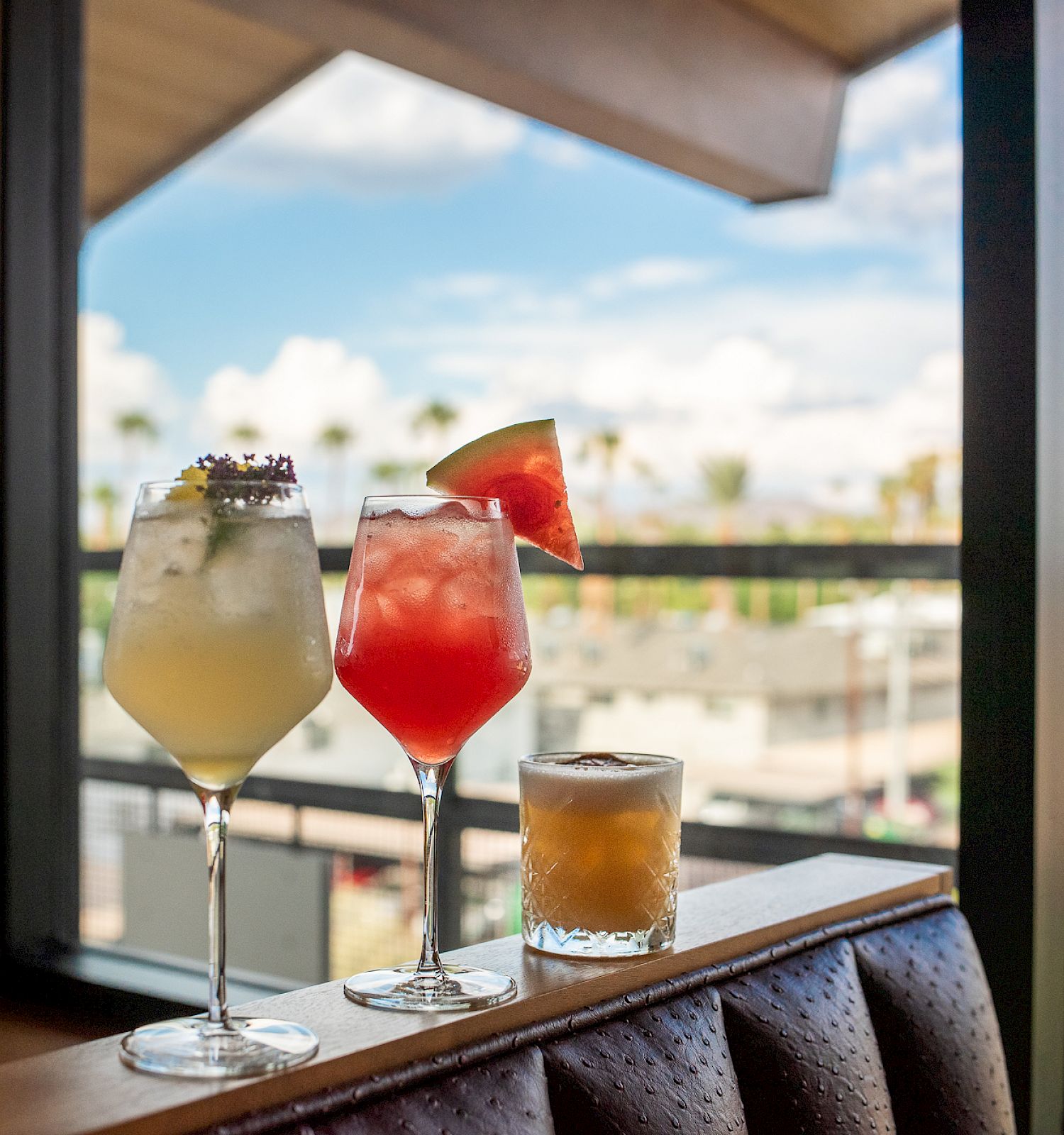 Three cocktails are showcased, each in different glasses, on a ledge with a scenic, sunny view outside the window in the background.