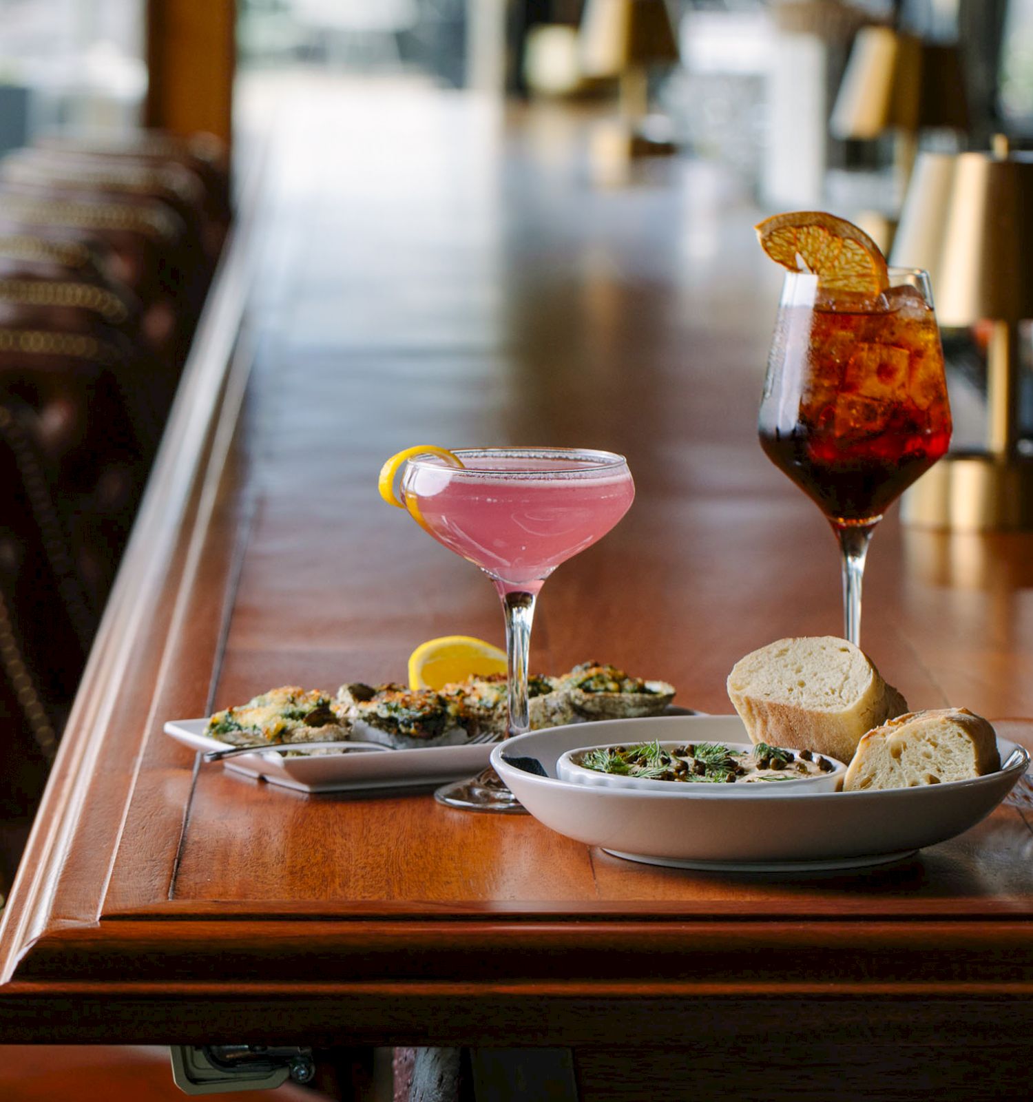 A bar counter with appetizers, bread, a pink cocktail, a tall drink with orange garnish, and a dish with olives in focus, leather chairs in the background.