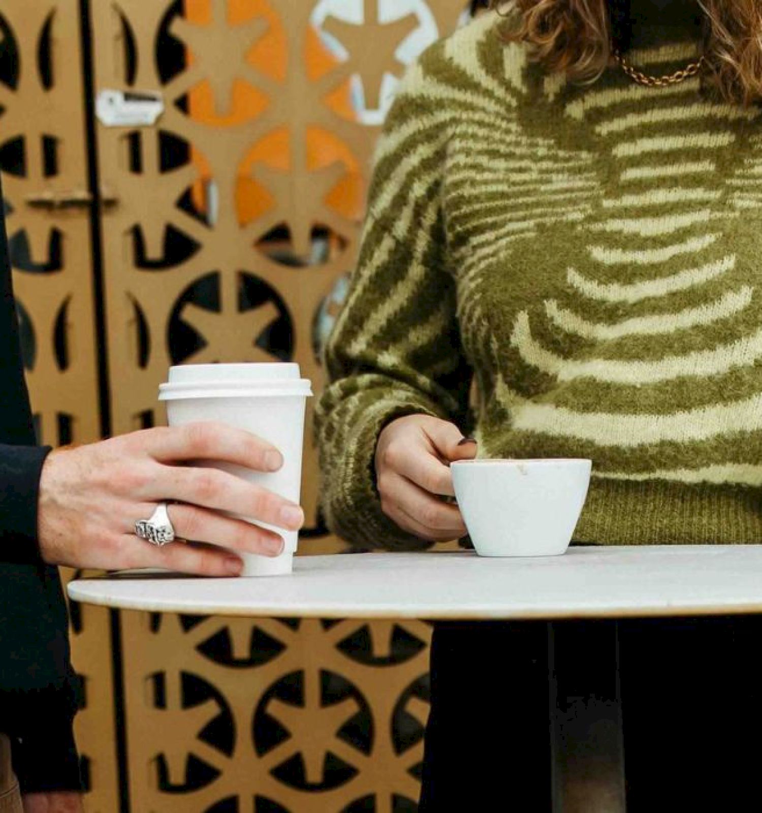 Two people at a table, one with a to-go coffee cup, the other with a small cup, wearing casual clothing.