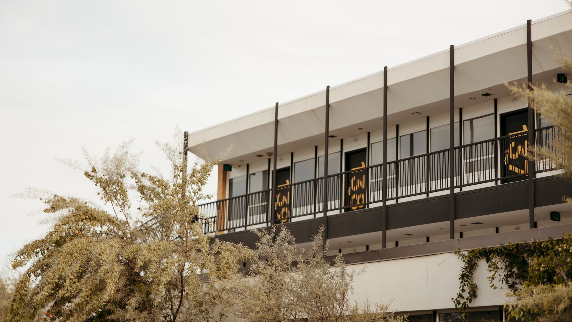 The image shows a modern two-story white building with black accents and balconies, and some greenery in the foreground.