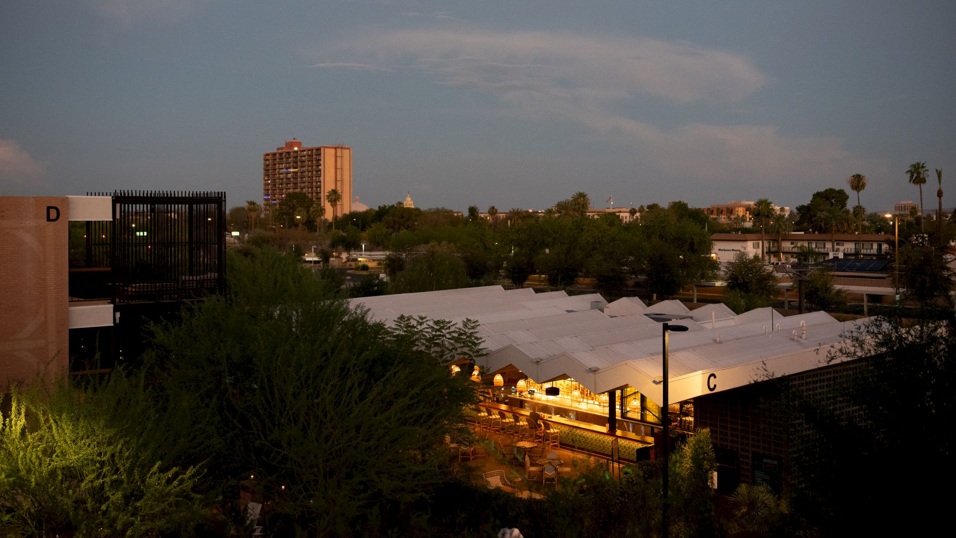 The image shows a cityscape at dusk with a mix of buildings, trees, and a lit-up market or event tent in the foreground.