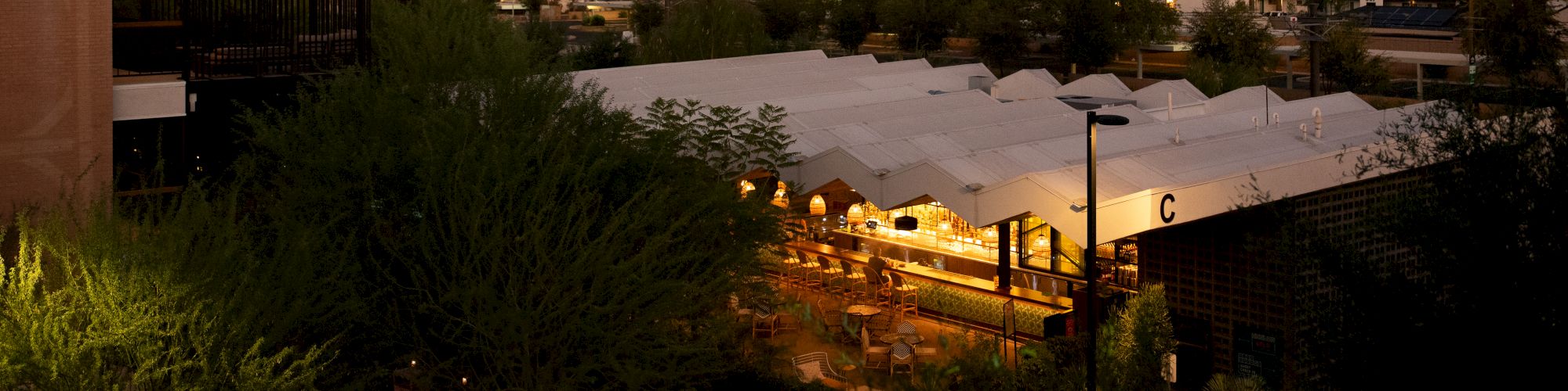 The image shows a cityscape at dusk with a mix of buildings, trees, and a lit-up market or event tent in the foreground.