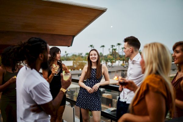 A group of people is gathered on a rooftop having a conversation, with drinks in hand and smiling faces, under an evening sky.