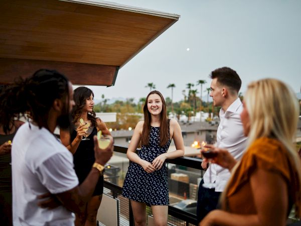 A group of people is gathered on a rooftop having a conversation, with drinks in hand and smiling faces, under an evening sky.