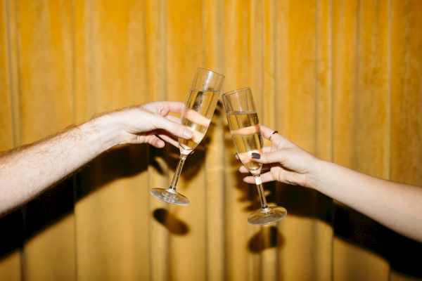 Two hands clinking champagne glasses against a golden, curtain-like background, suggesting a celebratory moment.