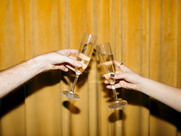 Two hands clinking champagne glasses against a golden, curtain-like background, suggesting a celebratory moment.