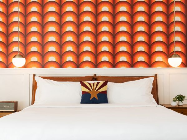 A modern bedroom with a patterned background, double bed, vibrant cushion, bedside tables, a retro radio, hanging lamps, and a small plant.