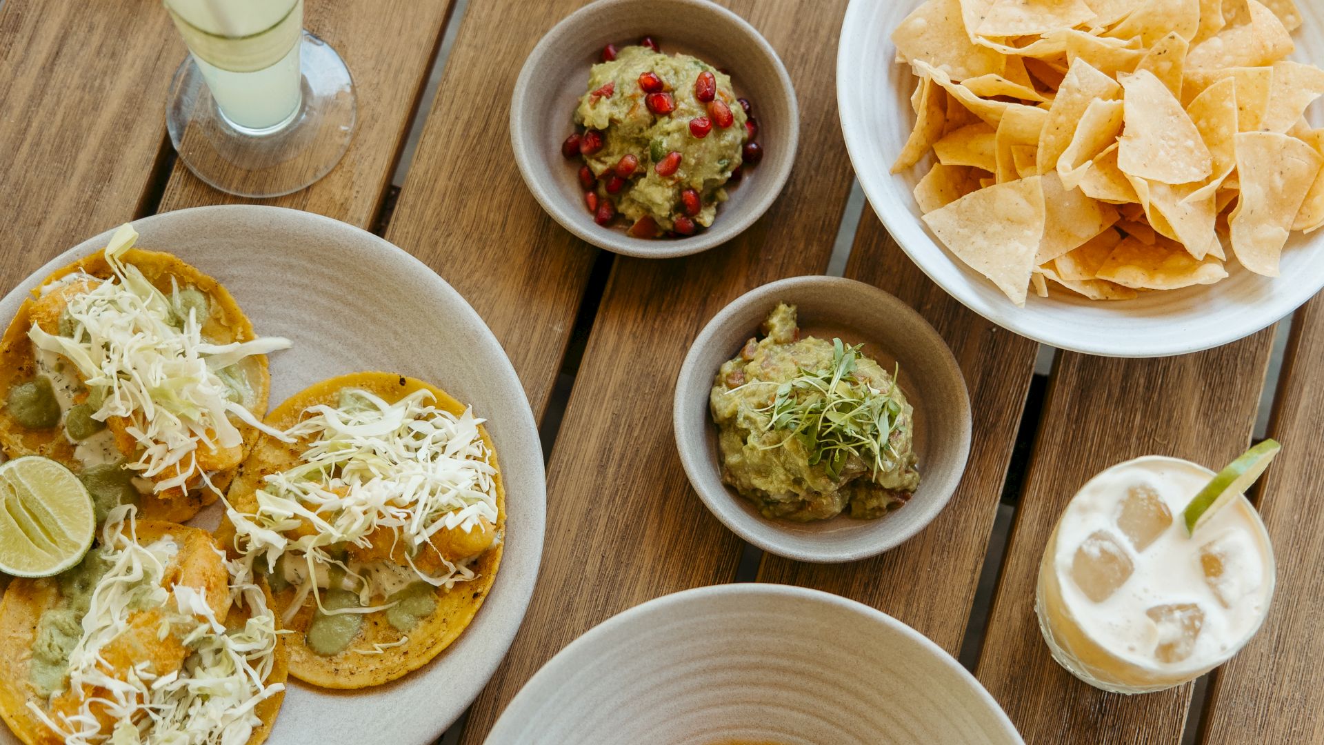 The image shows a table with assorted dishes including tacos, chips with dips, and various beverages. The table has a wooden texture.
