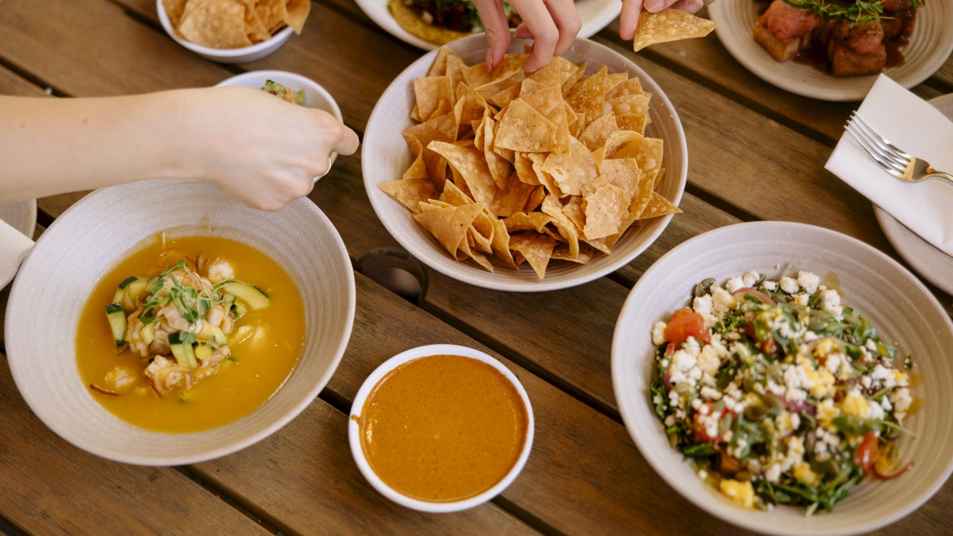 A shared table with tortilla chips, salsa, soup, and salads; people dip, scoop, and enjoy Mexican-inspired bowls and plates.