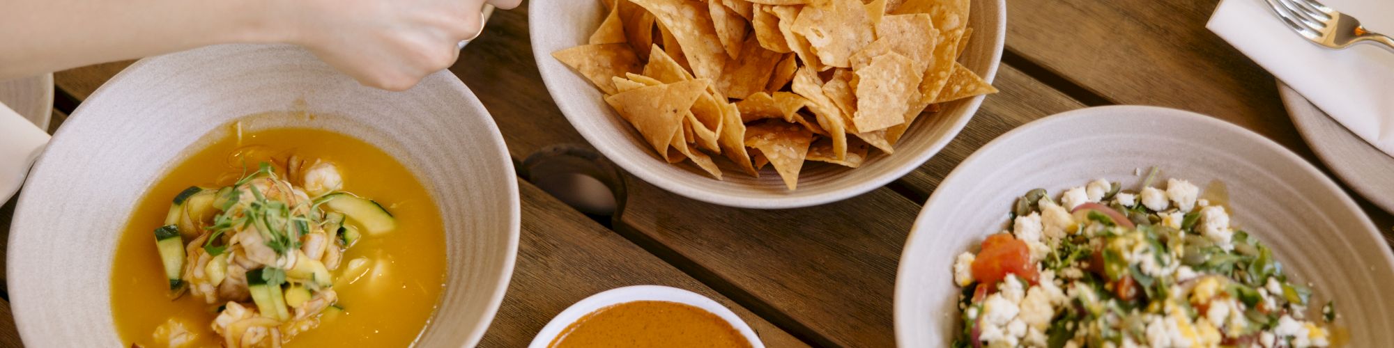 A shared table with tortilla chips, salsa, soup, and salads; people dip, scoop, and enjoy Mexican-inspired bowls and plates.