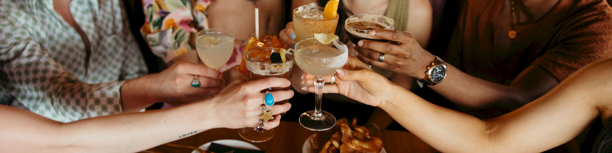 A group of friends is sitting around a table, toasting with cocktails and enjoying various snacks.