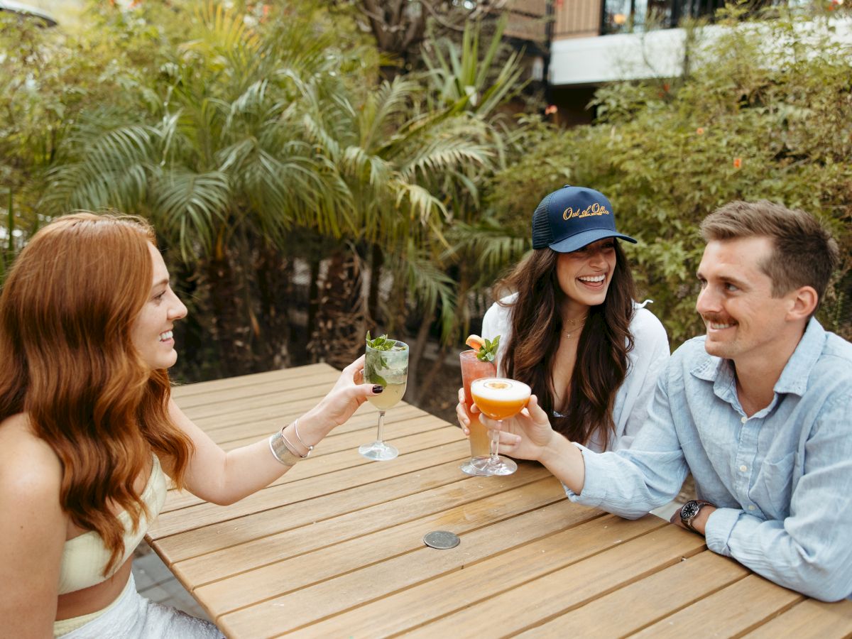 Three people sitting outside at a wooden table, holding drinks and smiling, surrounded by greenery.