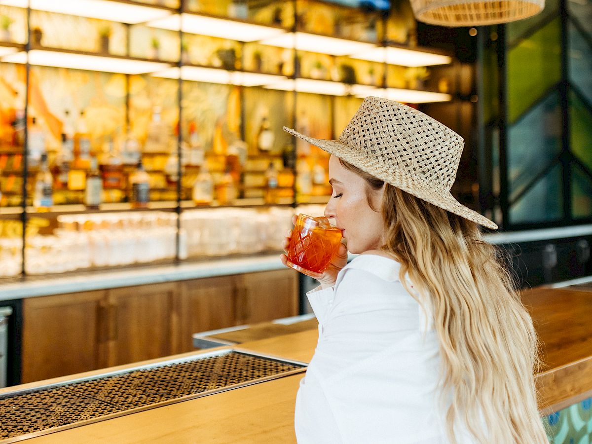 A person with long blonde hair, wearing a hat, is sipping a drink at a stylish bar with hanging lights and a TV in the background, ending the sentence.