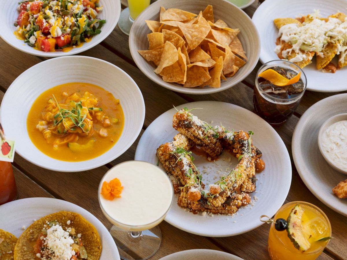 Assorted dishes and drinks including tacos, salad, chips, soup, corn, and cocktails arranged on a table.