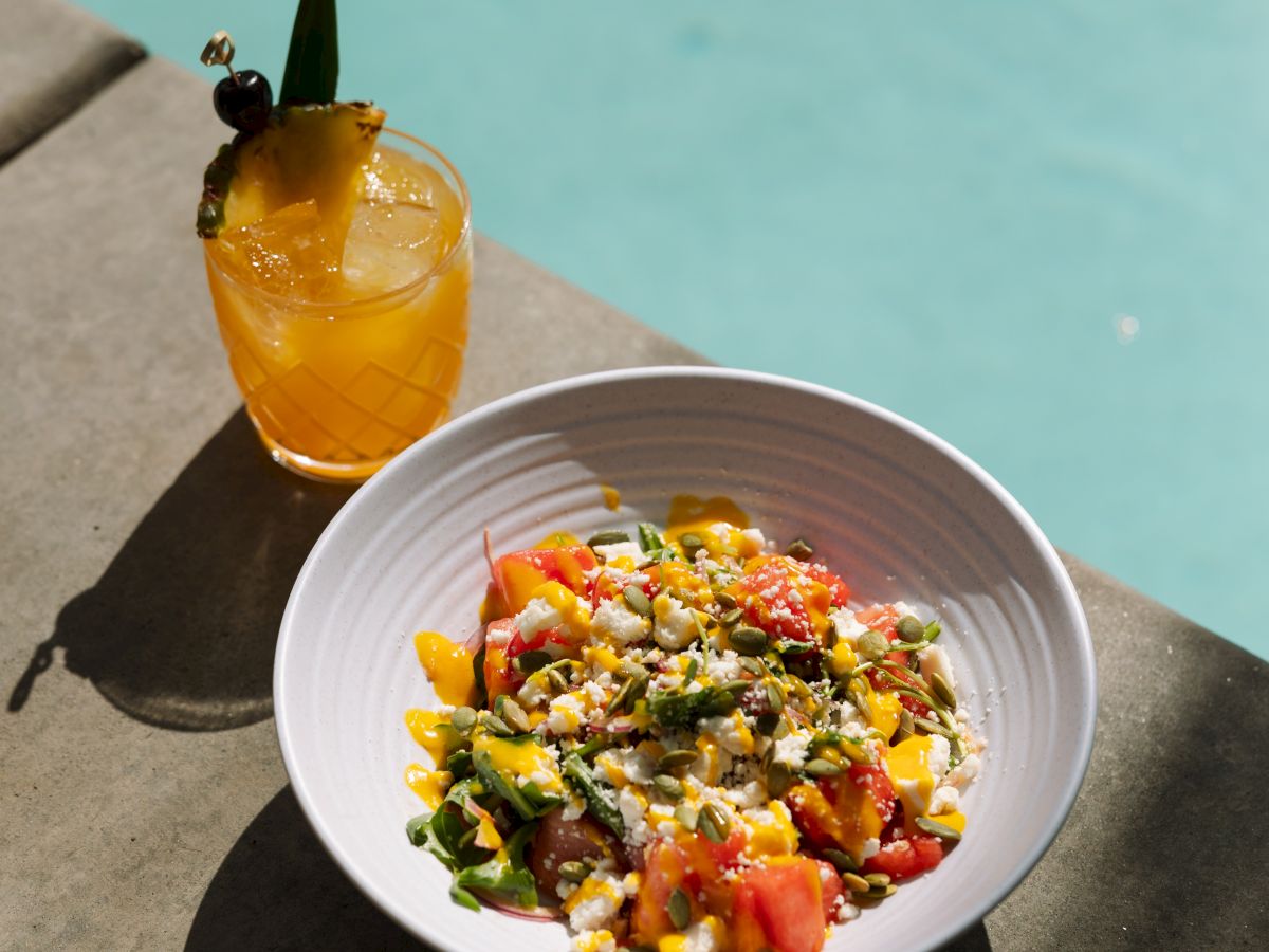 A refreshing fruit salad with greens and a citrus drink, both placed near a pool, creating a summery, relaxing vibe by the water.