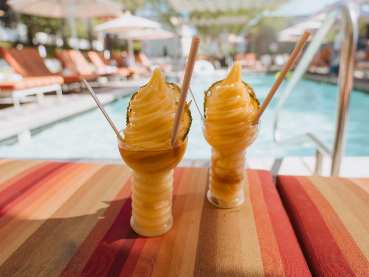 Two pineapple soft serve drinks with wooden straws by a poolside setup, surrounded by lounge chairs and umbrellas in the background.