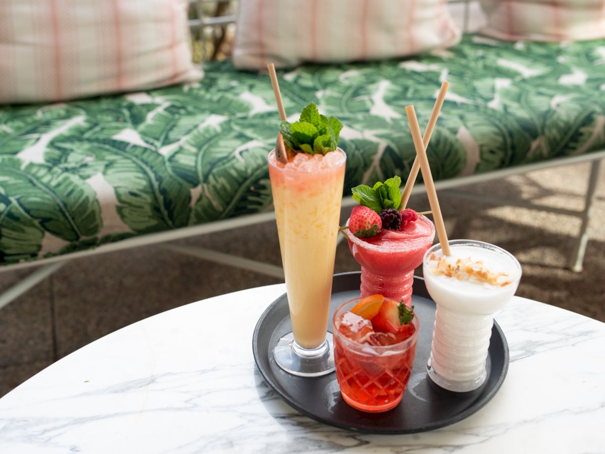 Assorted colorful drinks on a tray atop a marble table with a leafy green cushioned bench in the background.