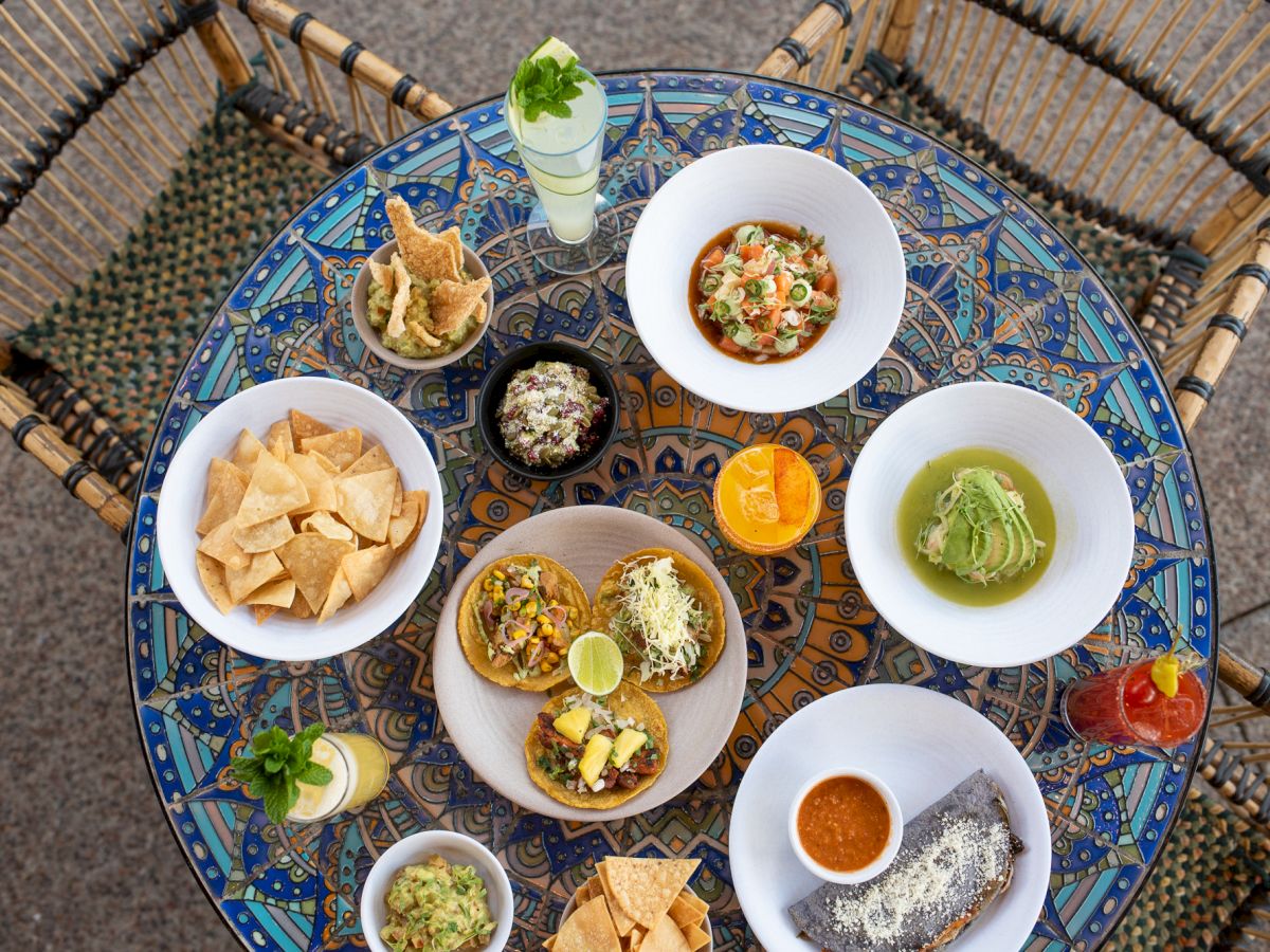 An overhead view of a table filled with a variety of Mexican dishes, chips, and drinks on a colorful, patterned tablecloth, surrounded by two chairs.