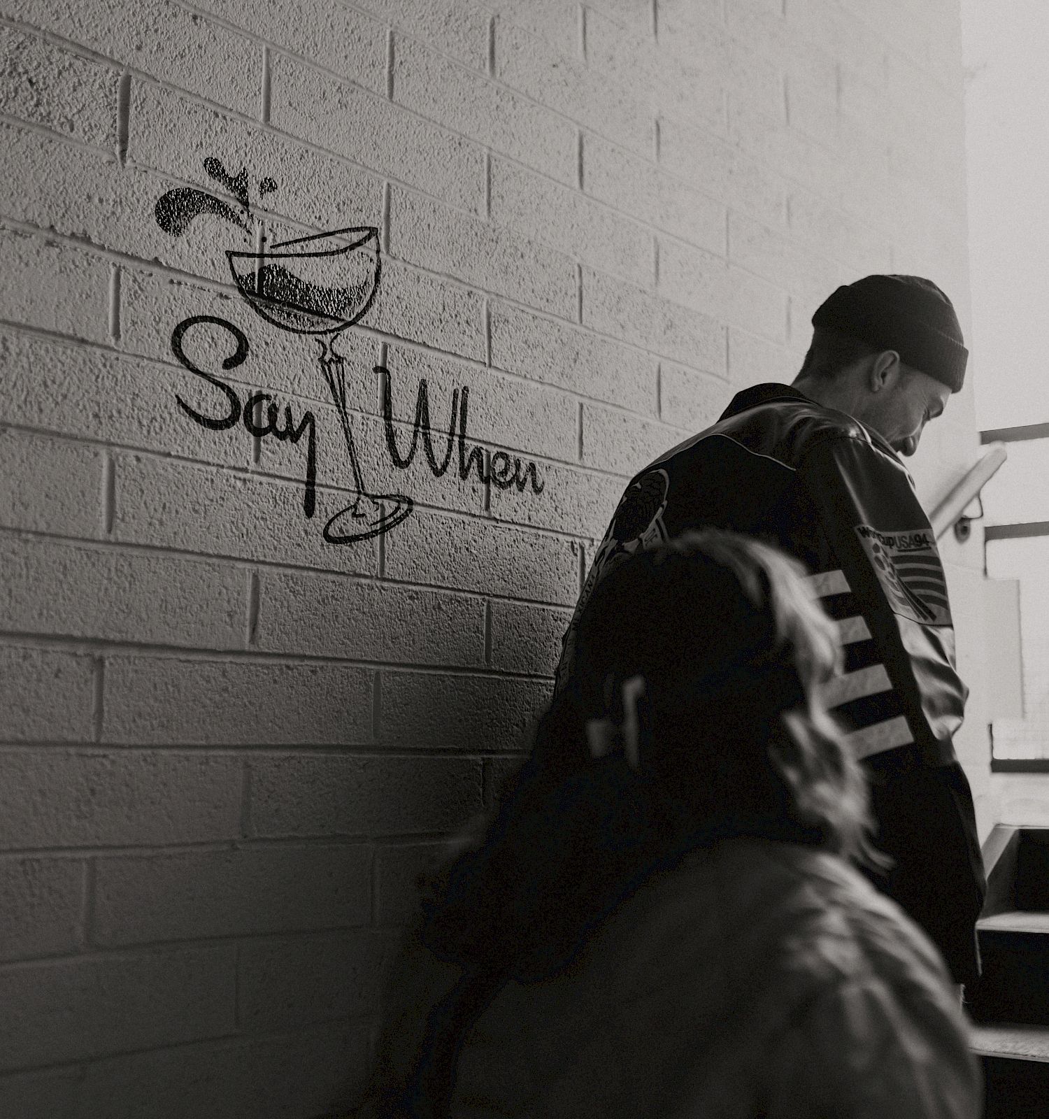 Two people are going up a staircase near a wall with a drawing of a glass and the text "Say When." They are dressed casually.