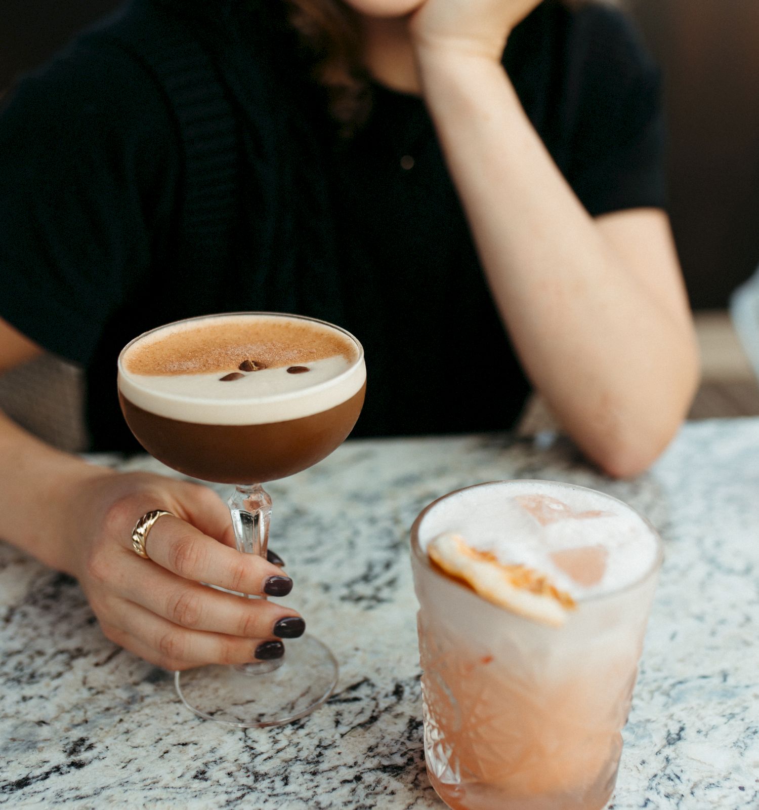 A person holding a glass of a creamy, coffee-like beverage is seated at a table with another drink nearby. The person is smiling gently.