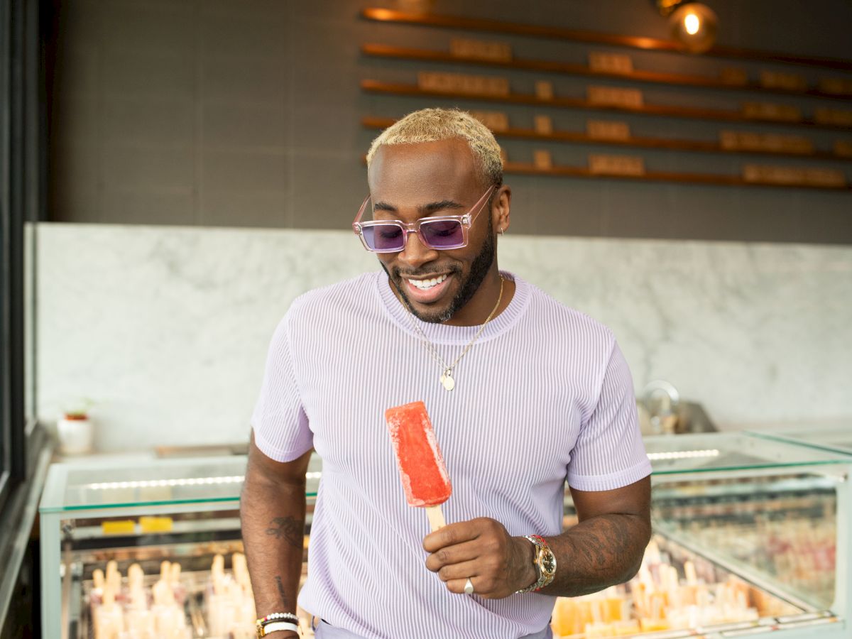 A man with blond hair and purple sunglasses is holding a red popsicle, smiling, and standing next to a counter with various popsicles inside.