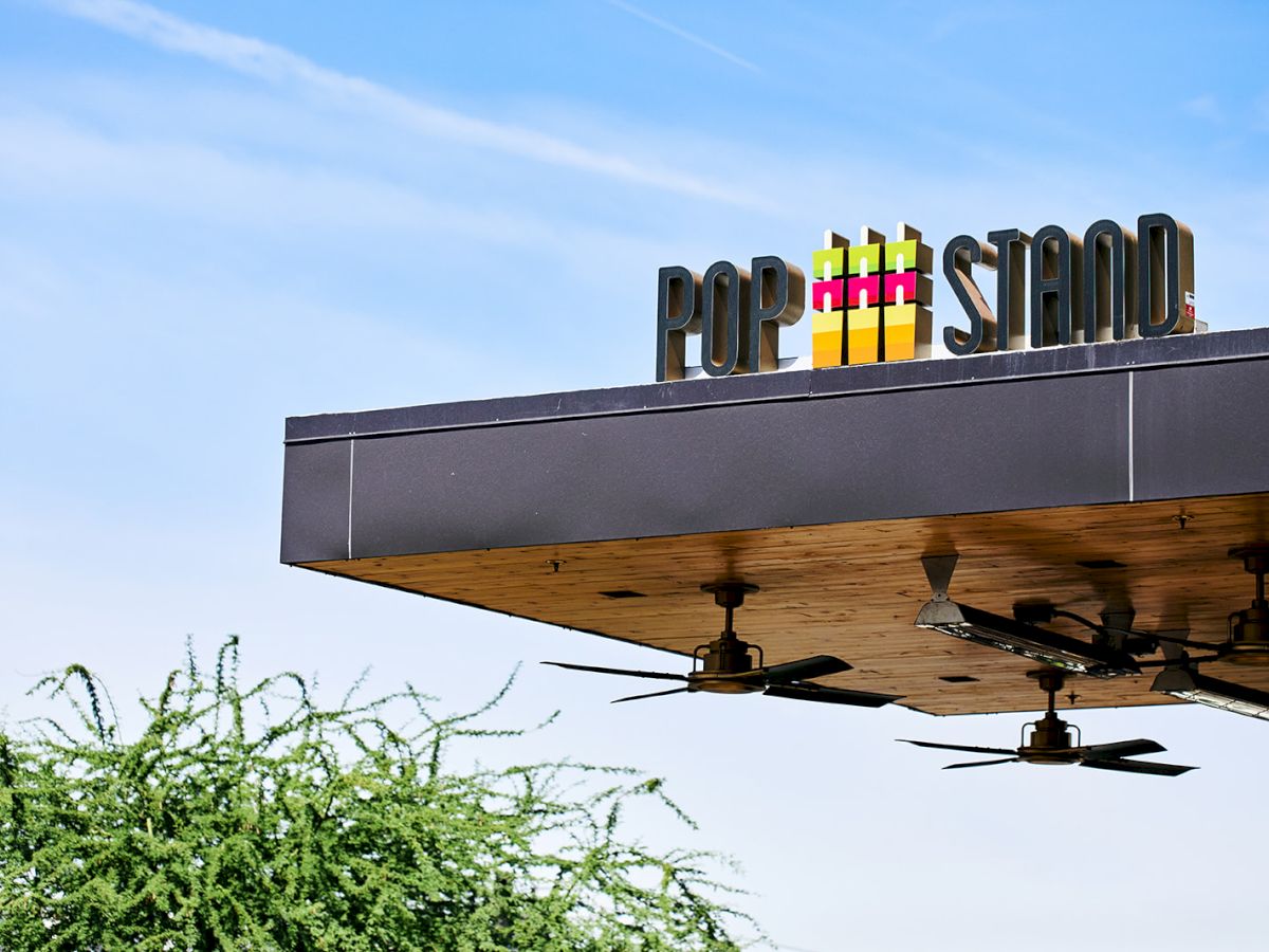 A sign atop a structure reads "POP STAND" against a blue sky. Ceiling fans are visible underneath the structure, and greenery is seen below.