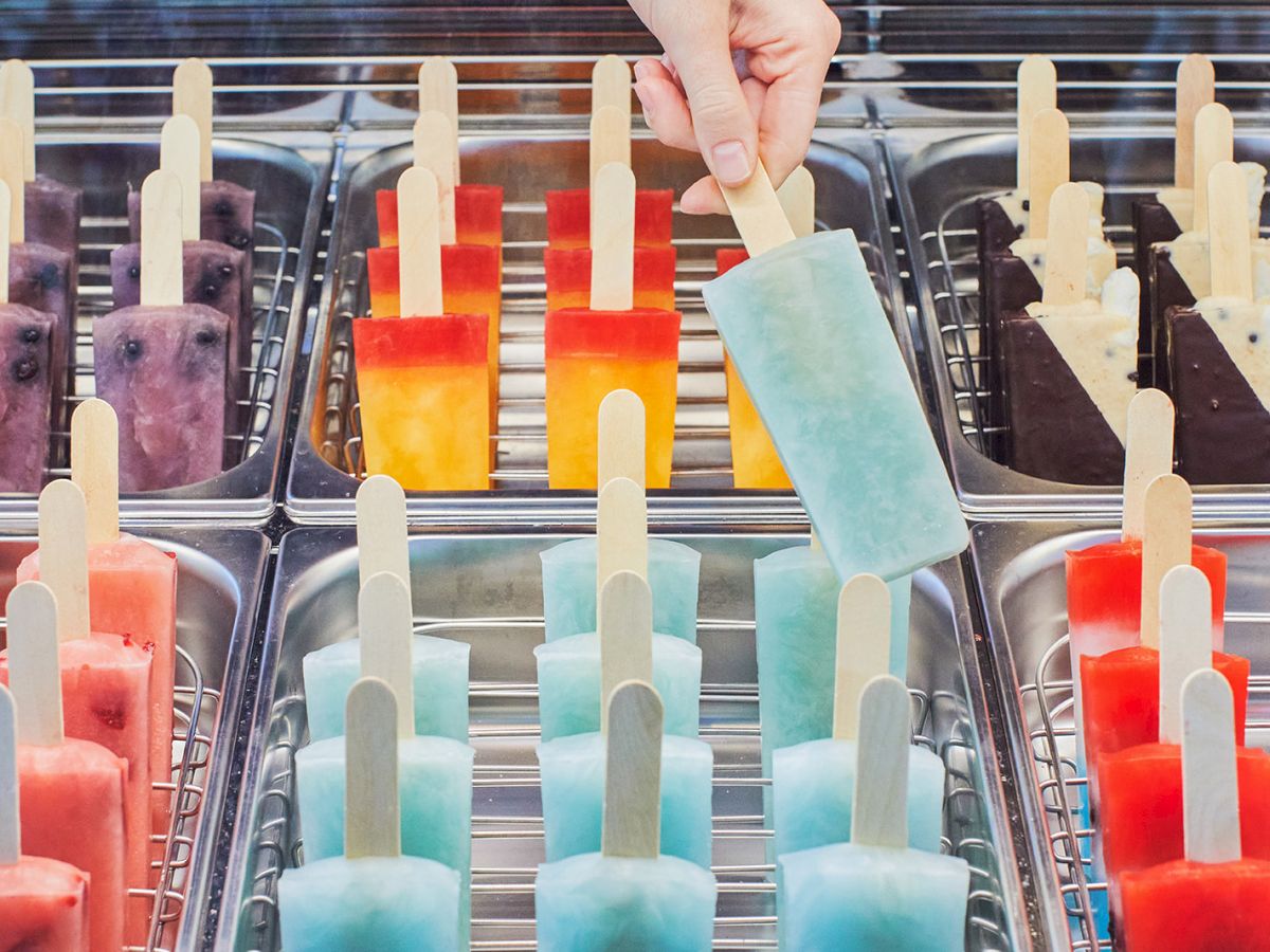 A variety of colorful ice pops are displayed in trays, with a hand picking up a blue one.