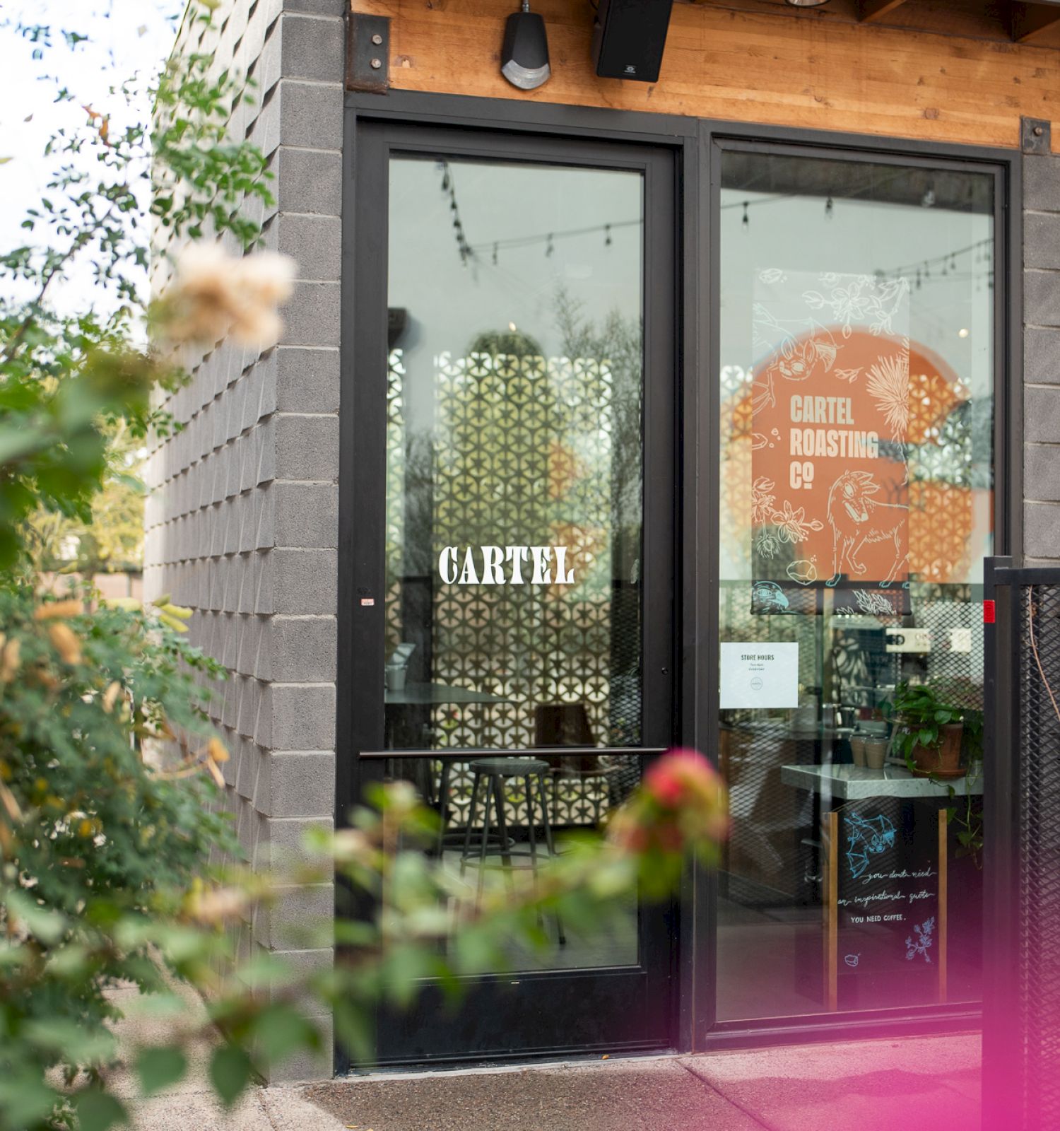 The image depicts the entrance to a café with the sign "CARTEL COFFEE LAB" on the door, surrounded by plants and a patio area.