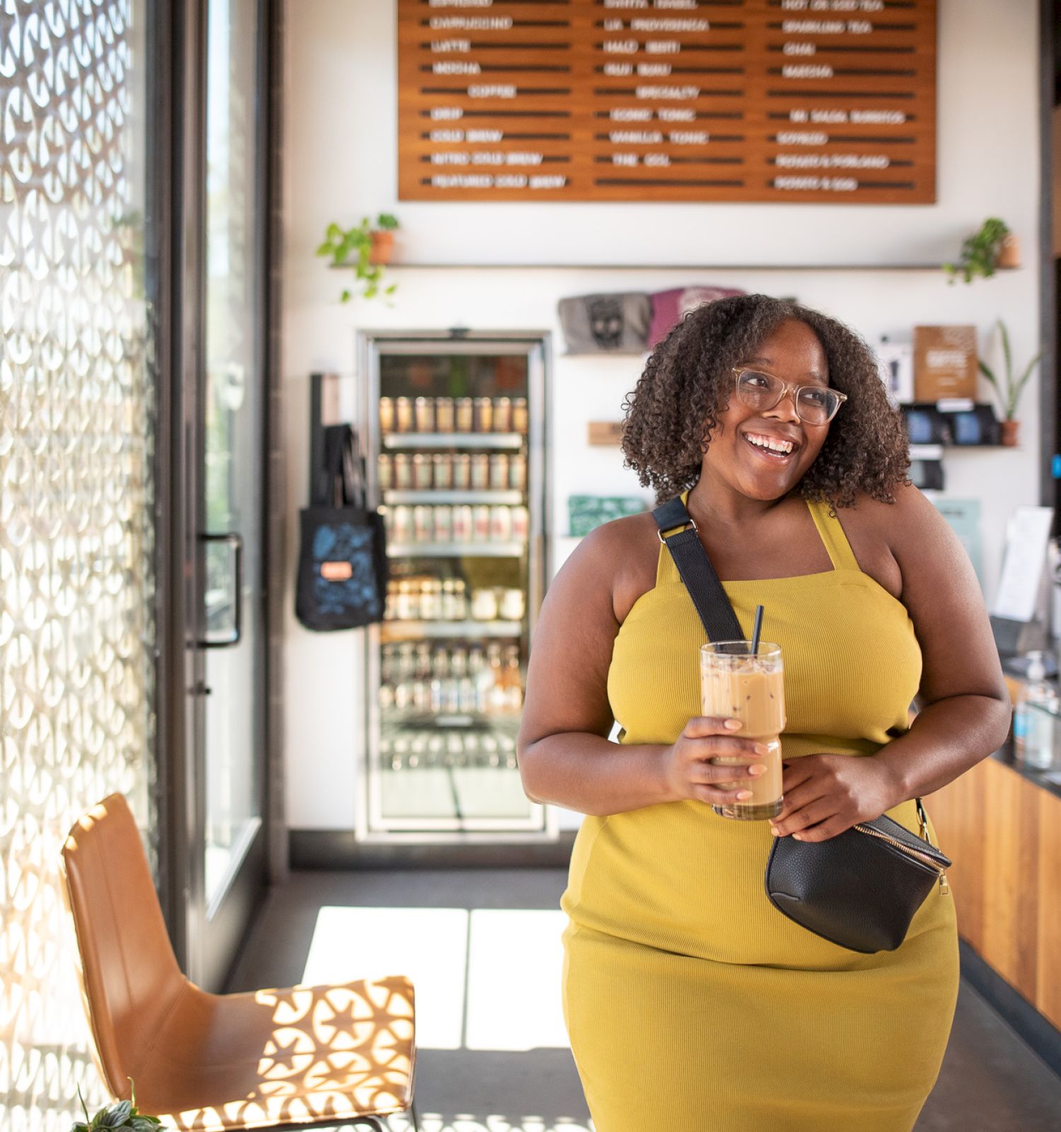 A person in a yellow dress is smiling, holding a drink, inside a café with a menu on the wall and a counter displaying baked goods.