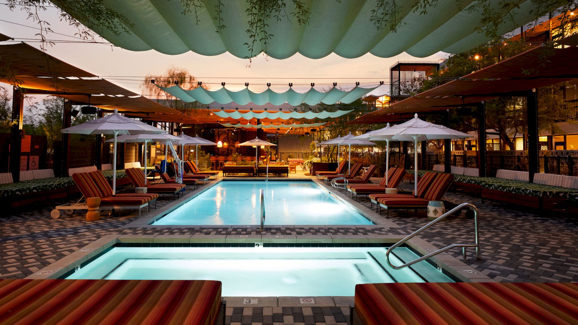 A luxurious outdoor pool area with lounge chairs, umbrellas, and overhead shade structures is shown. The ambiance is warm and inviting, perfect for relaxation.