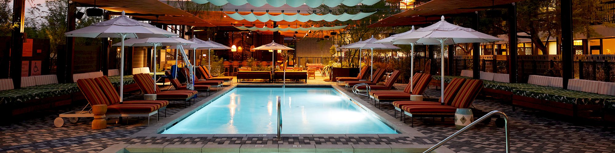 A luxurious outdoor pool area with lounge chairs, umbrellas, and overhead shade structures is shown. The ambiance is warm and inviting, perfect for relaxation.