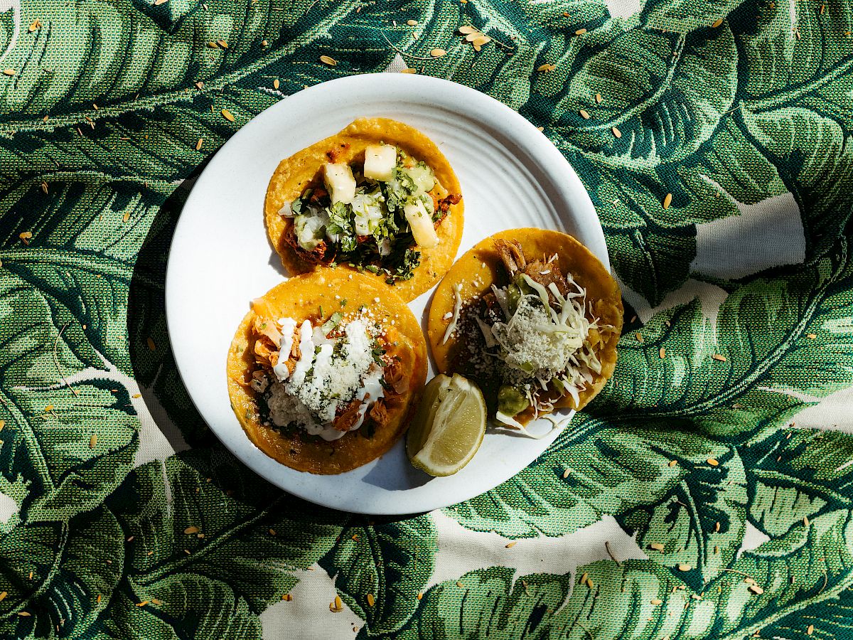 A white plate with three tacos and lime wedges sits on a green leaf-patterned fabric.