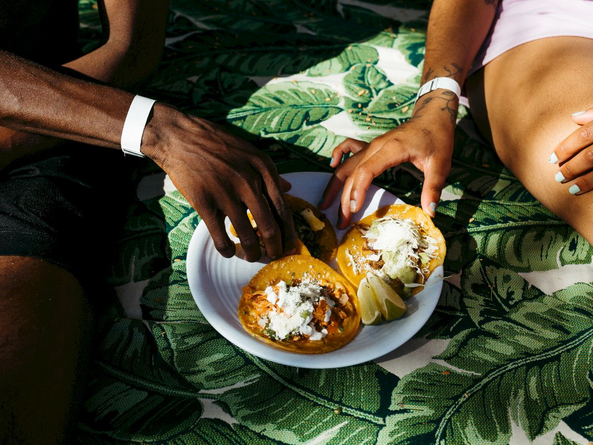 Two people are enjoying tacos outdoors, reaching for the food on a leaf-patterned blanket, with wristbands visible on their arms.