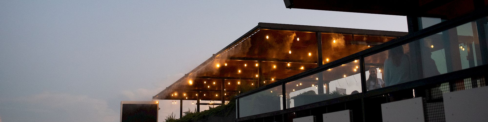 The image depicts a modern building with a rooftop terrace, adorned with string lights, overlooking a scenic view at dusk.