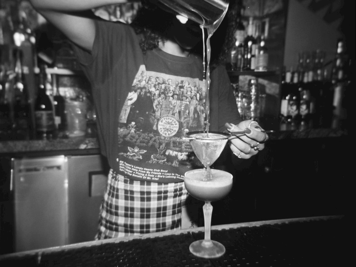 A person is pouring a drink into a glass at a bar, wearing a graphic T-shirt and an apron. The background shows shelves with various bottles.