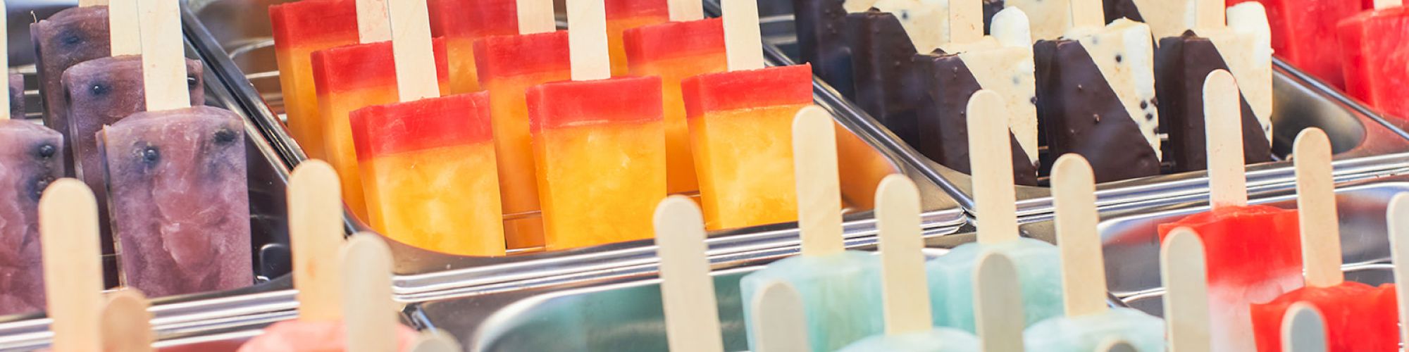 The image shows a variety of colorful popsicles arranged in metal trays, with flavors ranging from fruity to creamy.