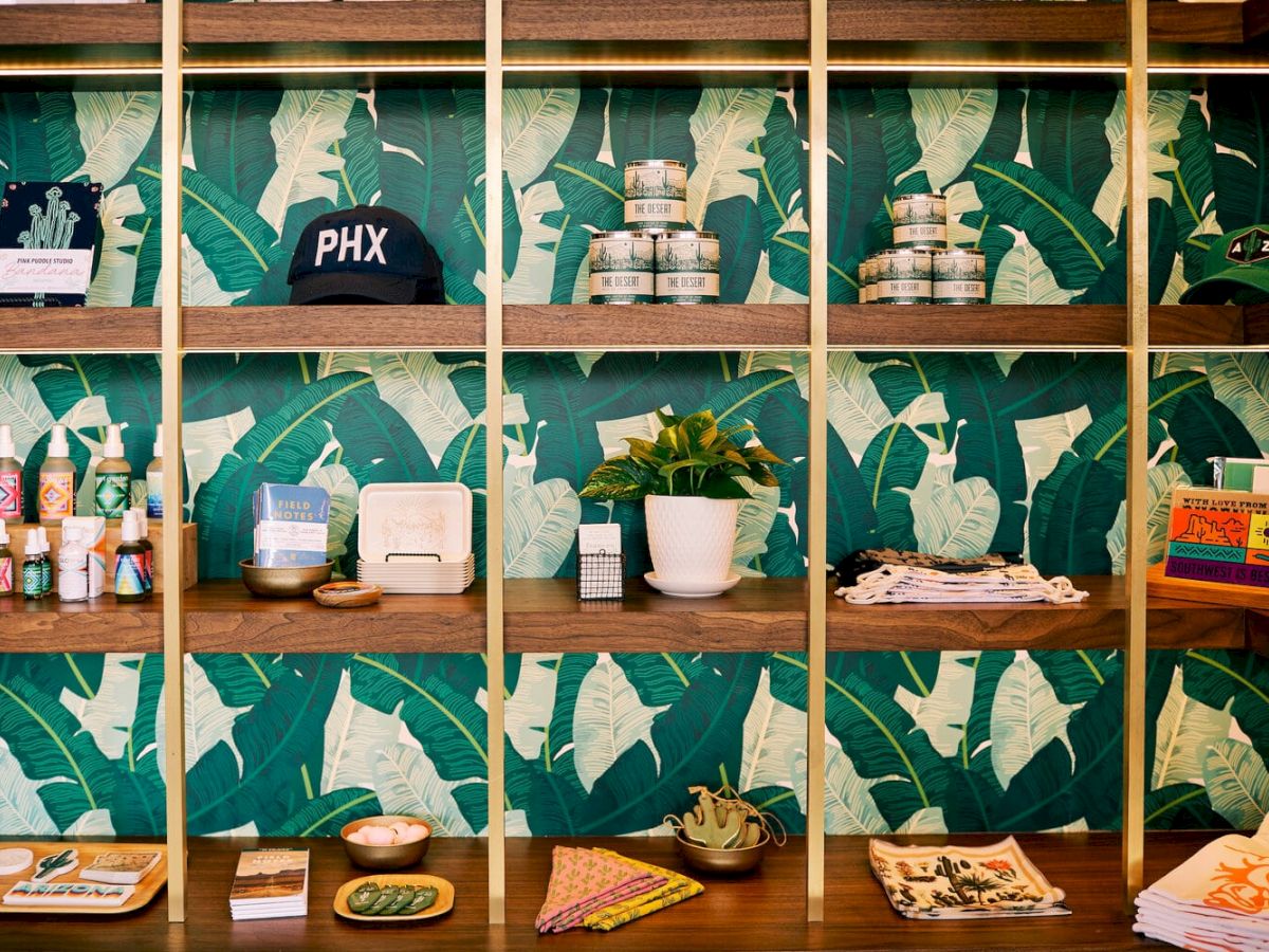 The image shows a wooden shelving unit with various items including a cap, candles, skincare products, a plant, and magazines against a leafy wallpaper.