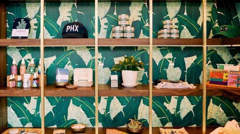 The image shows a wooden shelving unit with various items including a cap, candles, skincare products, a plant, and magazines against a leafy wallpaper.