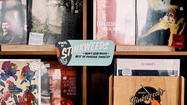 The image shows a collection of vinyl records displayed in shelves, including a sign that reads “STINKWEEDS” and a smaller wooden box with the same logo.