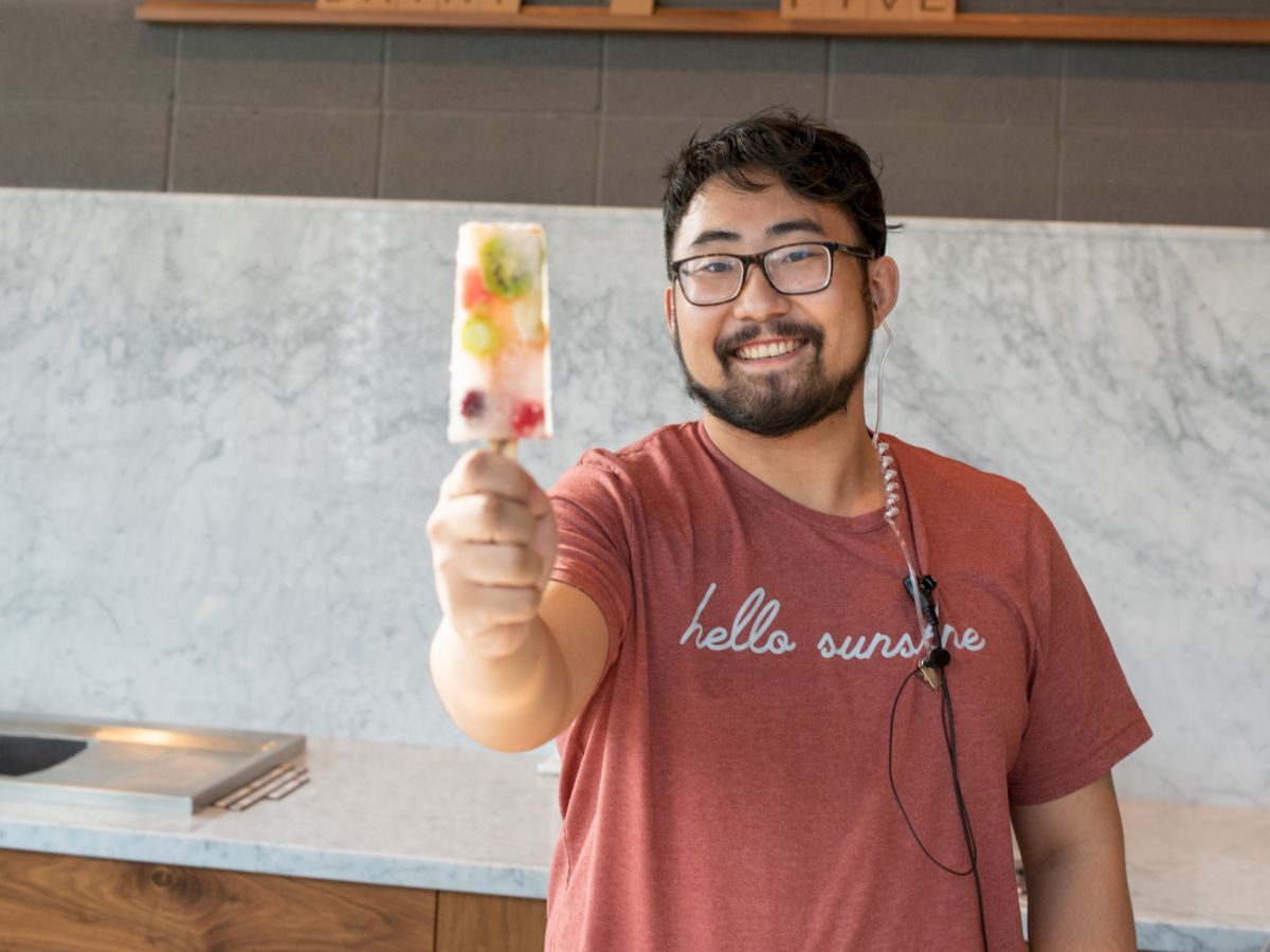 A smiling person, wearing a red "hello summer" t-shirt, is holding a colorful popsicle in a modern shop with a menu board on the wall behind them.